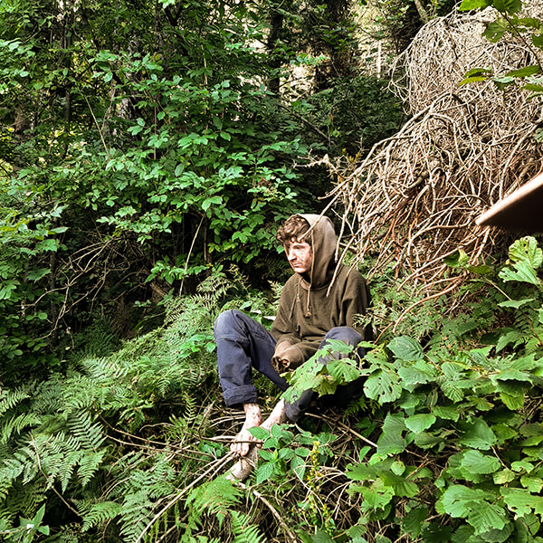 Ein Jugendlicher sitzt im Wald. Er trägt naturfarbene Kleidung.