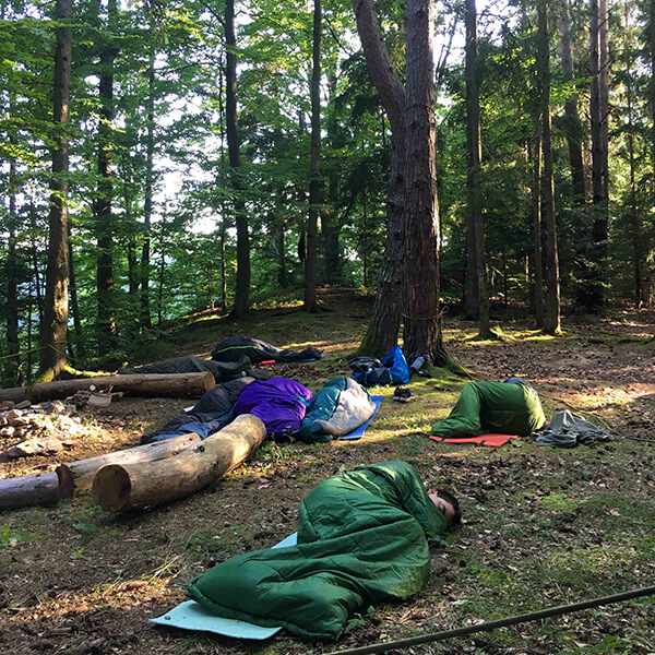 Jugendliche schlafen in Schläfsäcken auf Isomatten auf dem Waldboden.